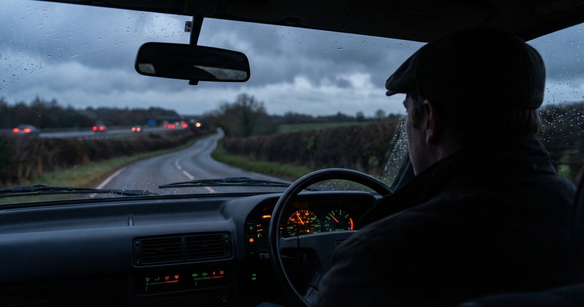 A driver at dusk on a dark UK road, dashboard glowing softly in the evening light